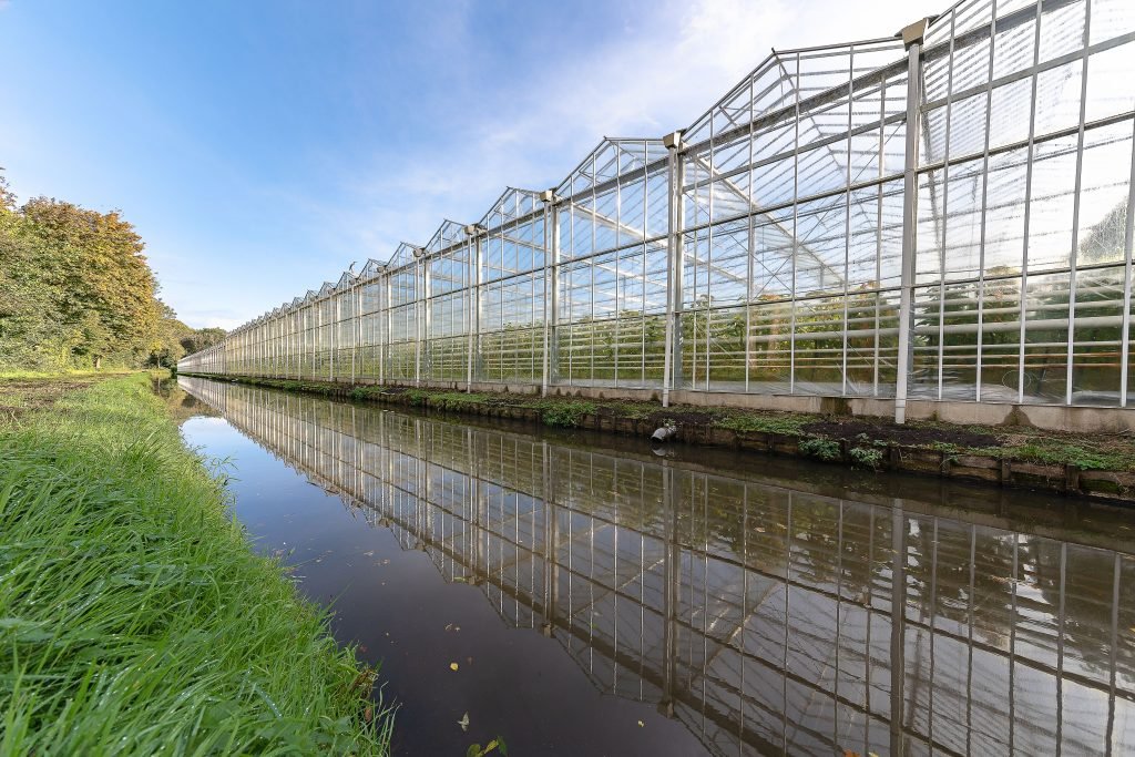 A large glass greenhouse with clear reflections on a calm canal, surrounded by greenery.